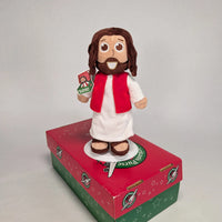 The Shoebox Jesus Doll standing on top of an Operation Christmas Child red and green shoebox.