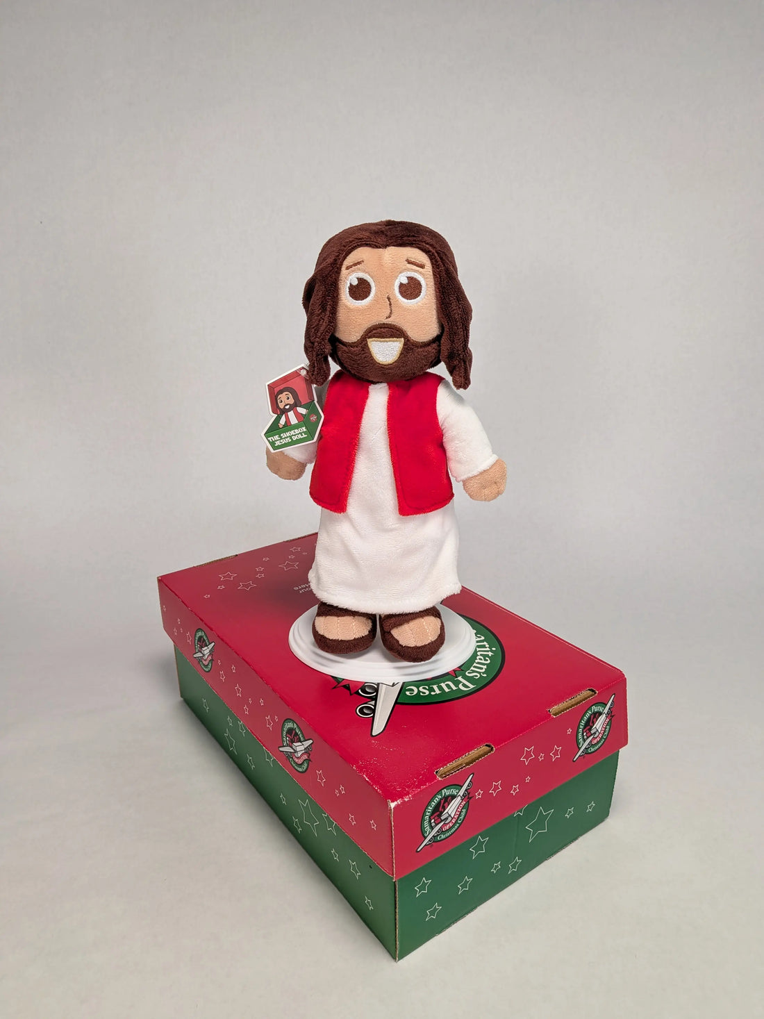 The Shoebox Jesus Doll standing on top of an Operation Christmas Child red and green shoebox.  